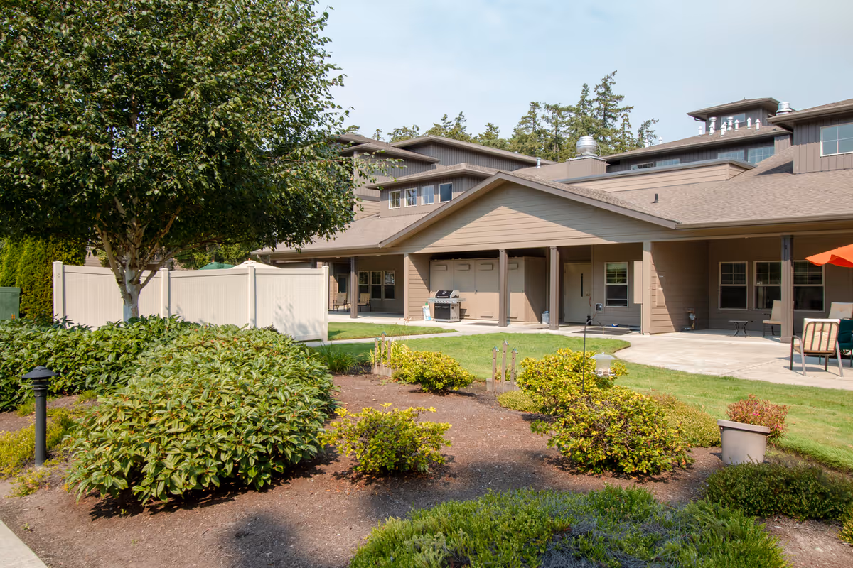 Outdoor view of Port Townsend Senior Living by Cogir showing a landscaped garden with bushes and a tree in the foreground, and a brown building with a covered patio area, outdoor seating, and a barbecue grill in the background under a partly cloudy sky.