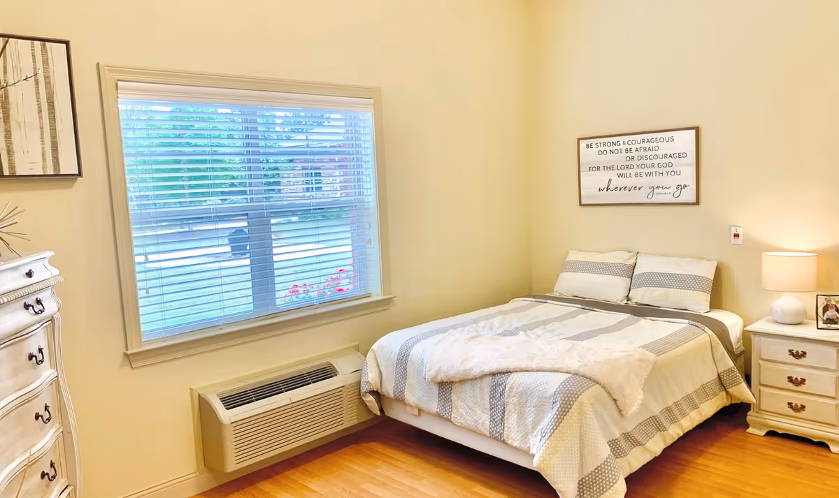 A cozy bedroom with a bed covered in a striped comforter and two pillows. There is a nightstand with a lamp and a framed photo on it to the right of the bed. Above the bed, a framed inspirational quote reads, 'Be strong & courageous do not be afraid or discouraged for the Lord your God will be with you wherever you go.' A window with blinds is on the left wall, and a white dresser with drawers is partially visible on the far left. The room has light-colored walls and wooden flooring.