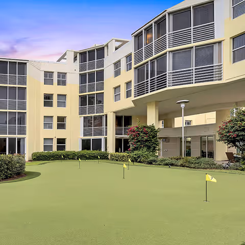 Outdoor putting green with several small yellow flags in front of a multi-story senior living facility building with large windows and balconies, surrounded by bushes and trees under a clear sky at sunset.