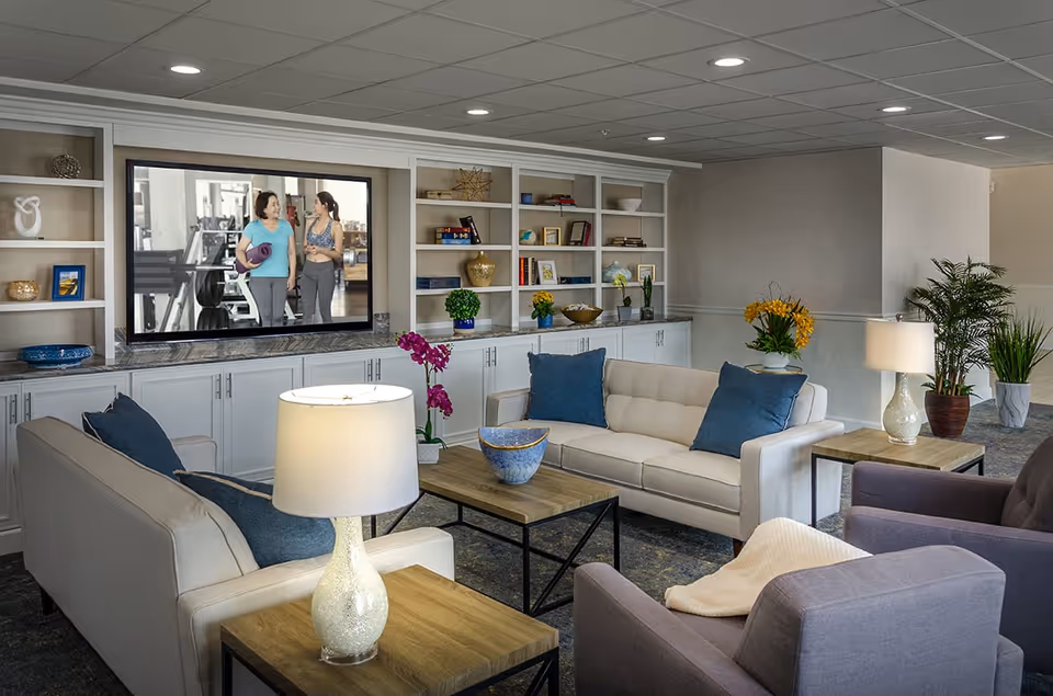 A bright communal living room with sofas, armchairs, coffee tables, built-in shelving and a wall-mounted TV showing two women in a gym.