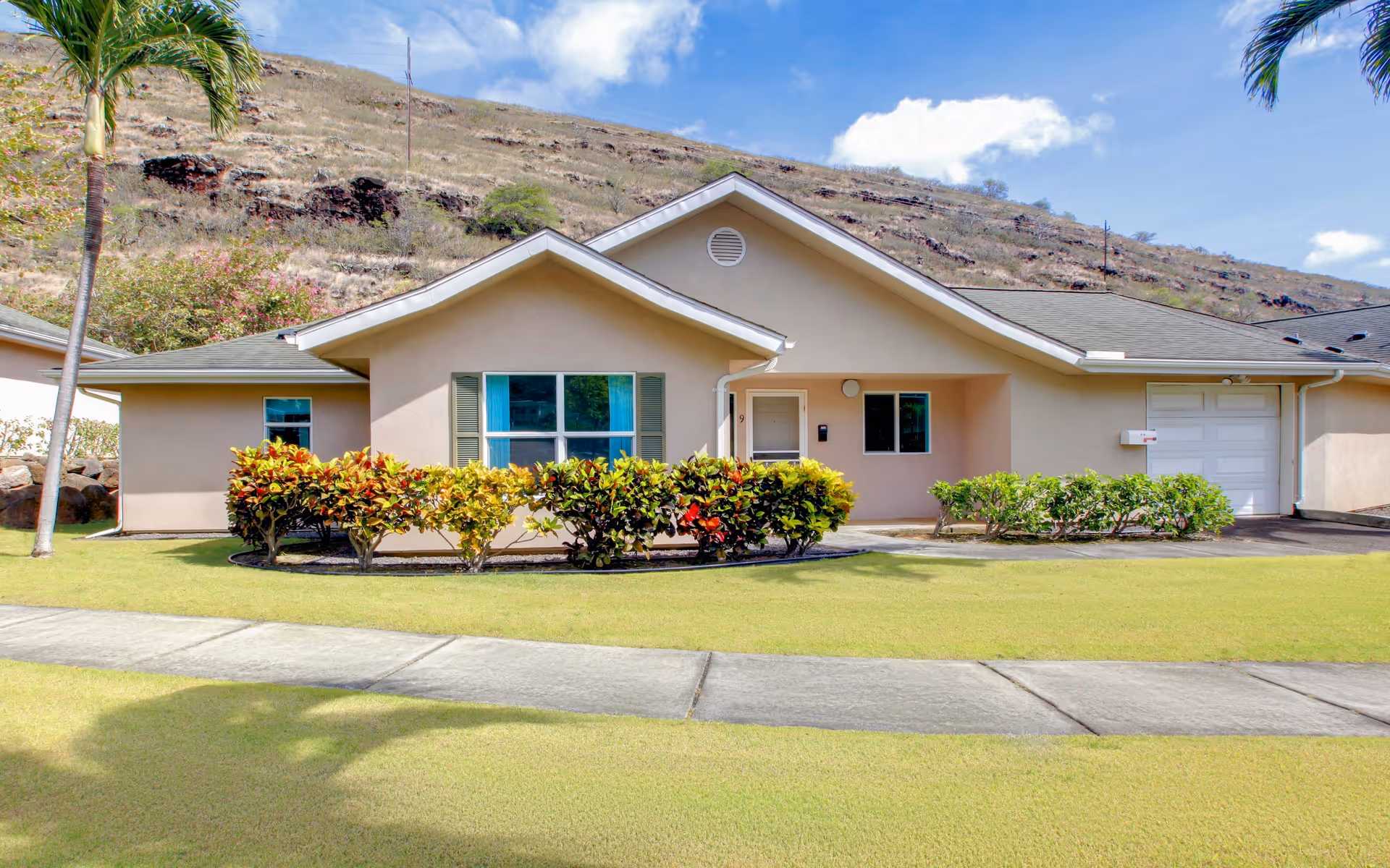 Single-story beige residential building with a sloped roof, front door, windows with shutters, and a garage. The building is surrounded by a well-maintained lawn, colorful bushes, palm trees, and a sidewalk in front. A rocky hillside is visible in the background under a partly cloudy sky.