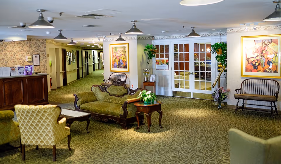 Carpeted senior living lounge with vintage sofas, armchairs, side tables, framed artwork, and double glass doors.