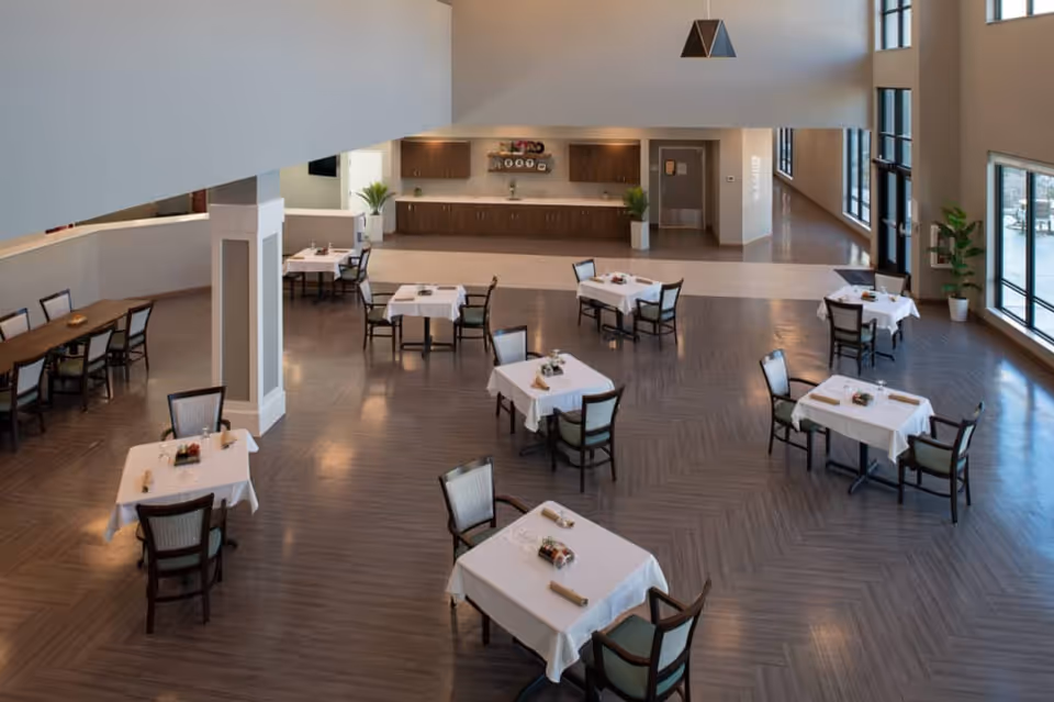 Spacious dining room with multiple tables covered in white tablecloths, each set with chairs and small centerpieces. Large windows on the right side allow natural light to fill the room. The back wall features wooden cabinets and a countertop with plants on either side. The floor has a wood pattern, and the ceiling is high with modern lighting.