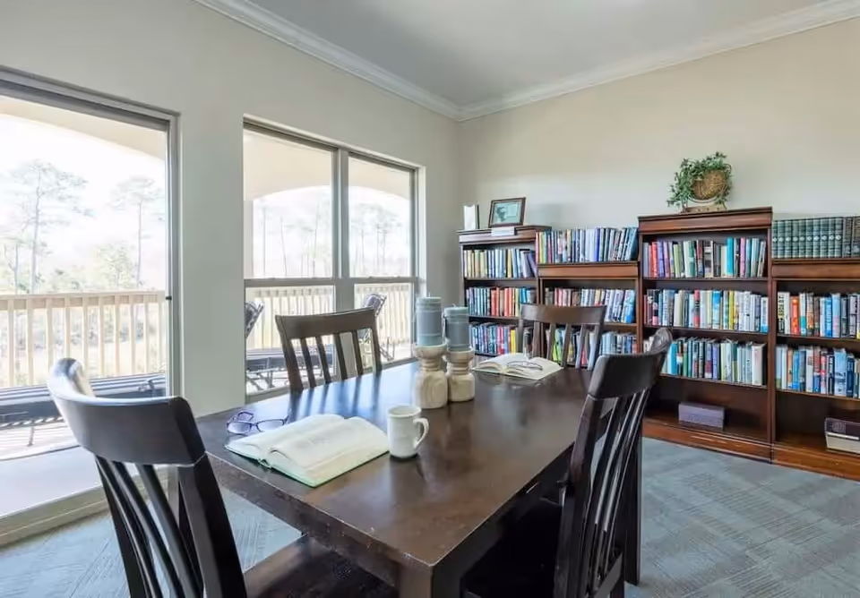 A bright room with large windows overlooking an outdoor area with trees. Inside, there is a dark wooden table with four chairs around it. On the table are two open books, a pair of glasses, a white coffee mug, and decorative candle holders. Against the wall are three wooden bookshelves filled with books and a small plant on top of one shelf.