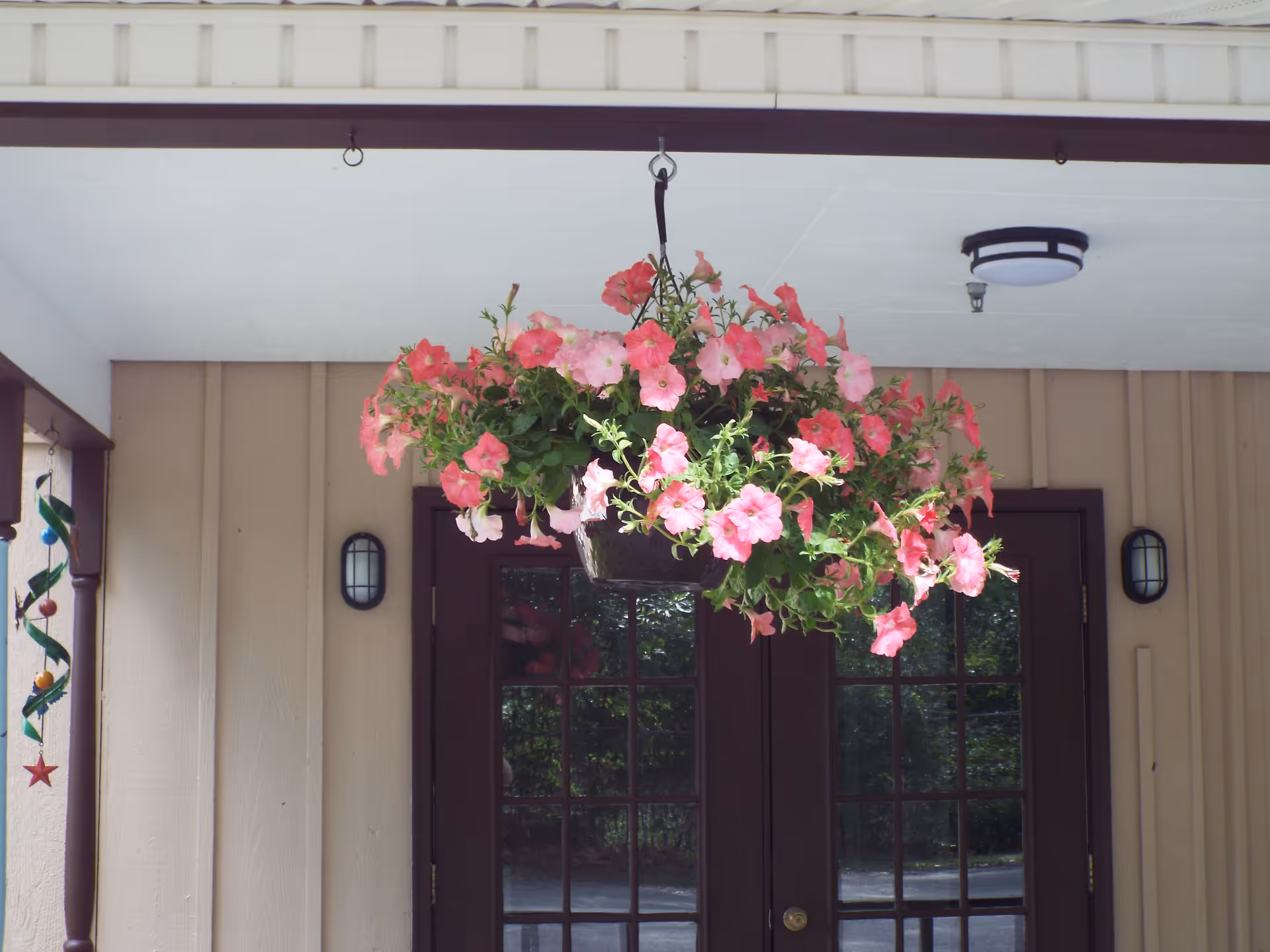A hanging basket filled with pink and light pink flowers is suspended from the ceiling of a covered porch area. Behind the flowers, there are double glass doors with dark brown frames, beige siding on the walls, two wall-mounted outdoor lights, and a ceiling light fixture.