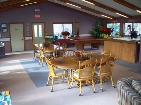 A spacious common area with wooden tables and chairs arranged in the center. The room has a high ceiling with exposed wooden beams and several windows letting in natural light. There is a pool table in the background, a counter area to the right, and some plants and decorations placed around the room.