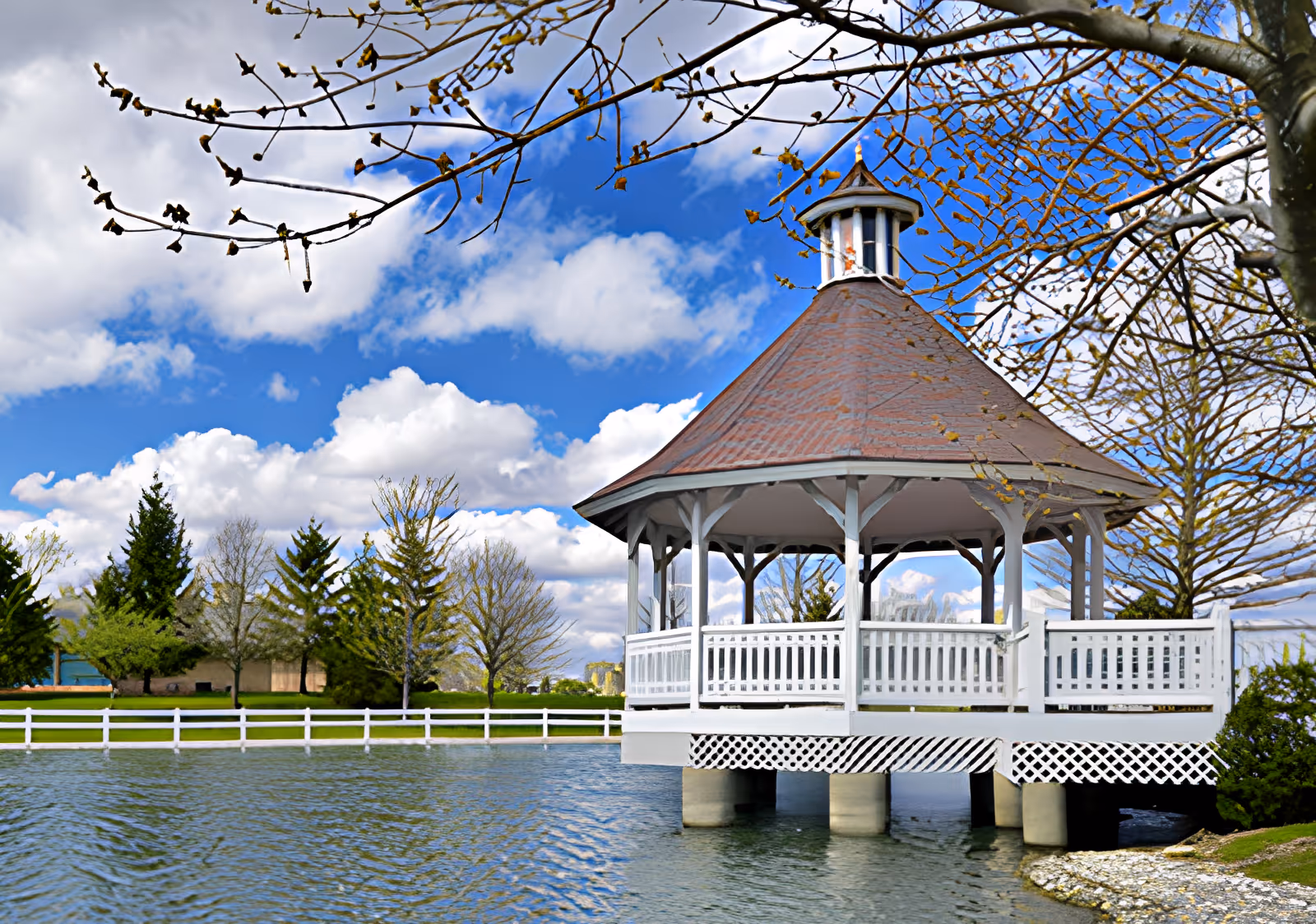 A white gazebo with a brown roof built on pillars over a pond, surrounded by trees and a white fence under a partly cloudy blue sky.