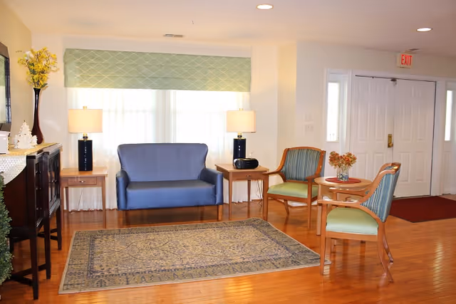 A bright seating area with a blue loveseat, wooden chairs, side tables and lamps on a hardwood floor near double entry doors.