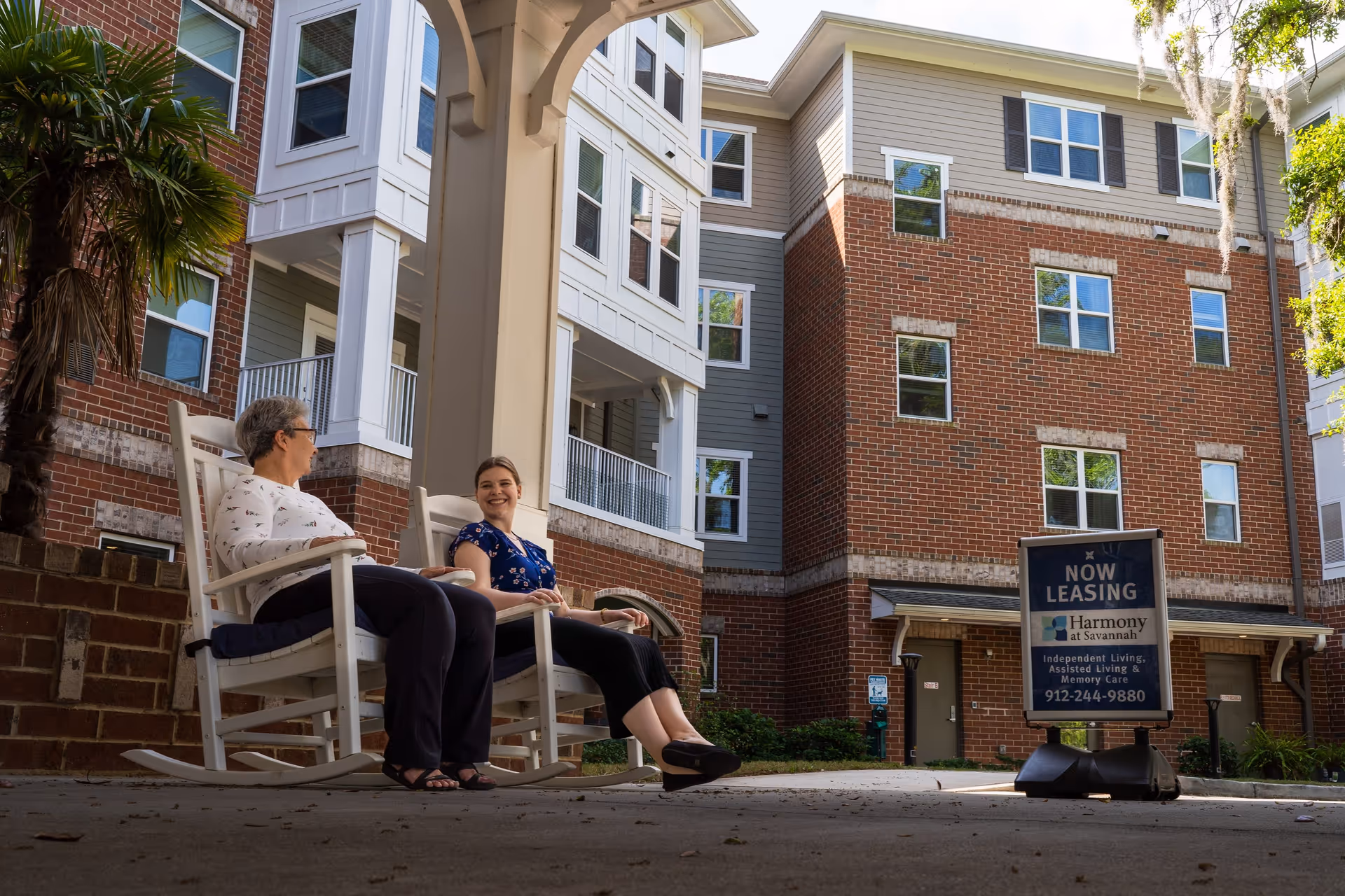 Two women sitting and talking on white rocking chairs under a covered outdoor area in front of a multi-story brick and siding building. A sign nearby reads 'Now Leasing Harmony at Savannah Independent Living, Assisted Living & Memory Care 912-244-9880'.