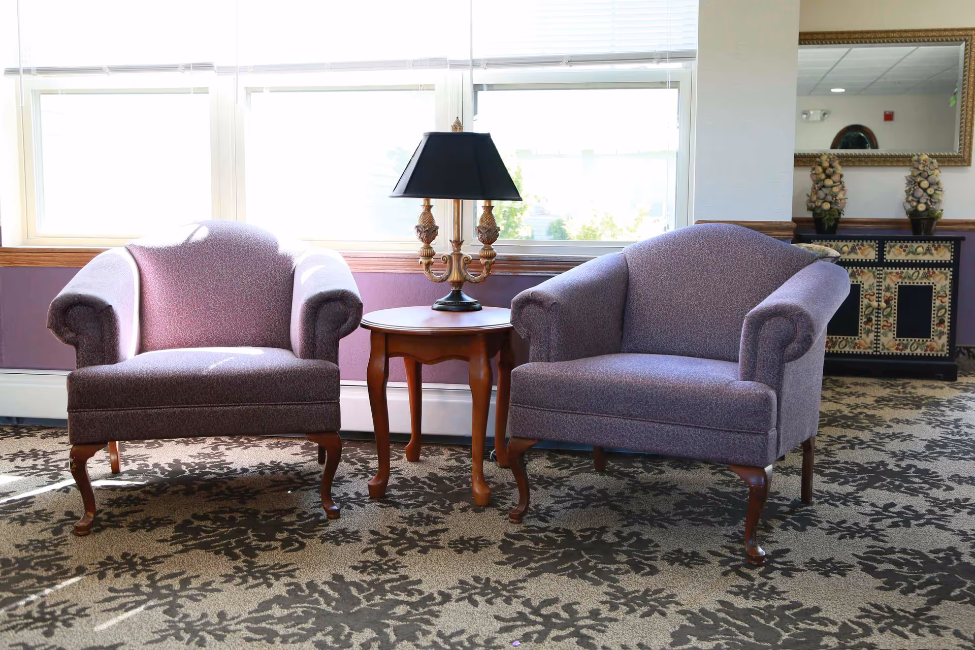 Two purple upholstered armchairs with wooden legs are positioned on either side of a small wooden side table with a decorative lamp on top. Behind the chairs is a large window letting in natural light. To the right, there is a decorative cabinet with a mirror above it and two ornamental pieces on top. The floor is carpeted with a floral pattern.