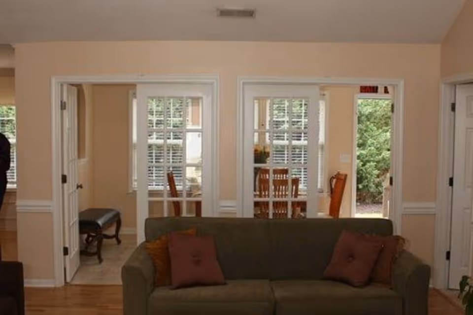 Interior view of a living room with a green sofa adorned with brown and red cushions. Behind the sofa, there are two sets of French doors leading to a dining area with wooden chairs and a table. The walls are painted beige with white trim, and there is a window with white blinds in the dining area. A door to the right leads outside to a garden area with greenery visible.