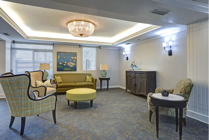 A well-lit living room area with a green sofa, two patterned armchairs, a round green ottoman, two side tables with lamps, a wooden cabinet, and a painting of water lilies on the wall. The room has a blue carpet, white walls, and a decorative ceiling light fixture.