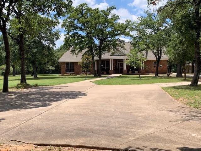 A single-story brick building with a covered front porch surrounded by trees and green grass under a partly cloudy sky. A wide concrete driveway leads up to the entrance.