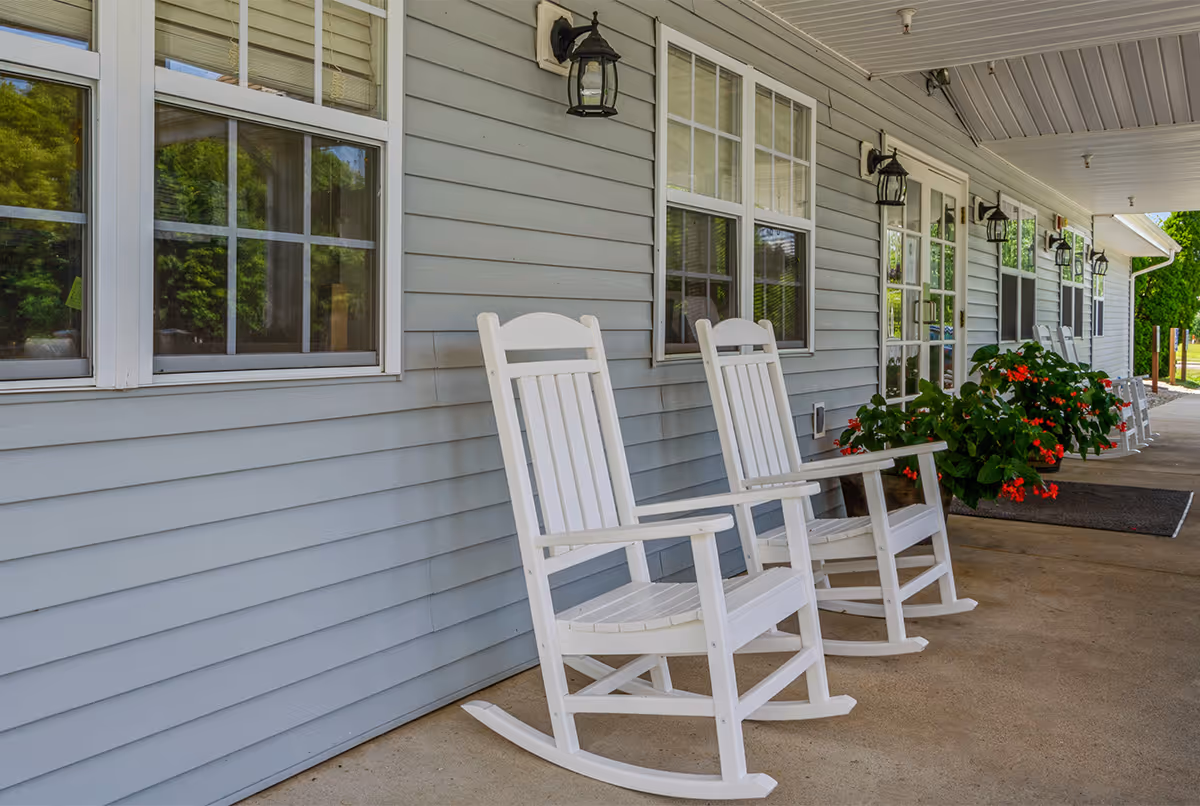 A covered porch area outside a building with light gray siding, featuring white rocking chairs and potted plants with red flowers. Several windows and outdoor wall lanterns are visible along the wall.