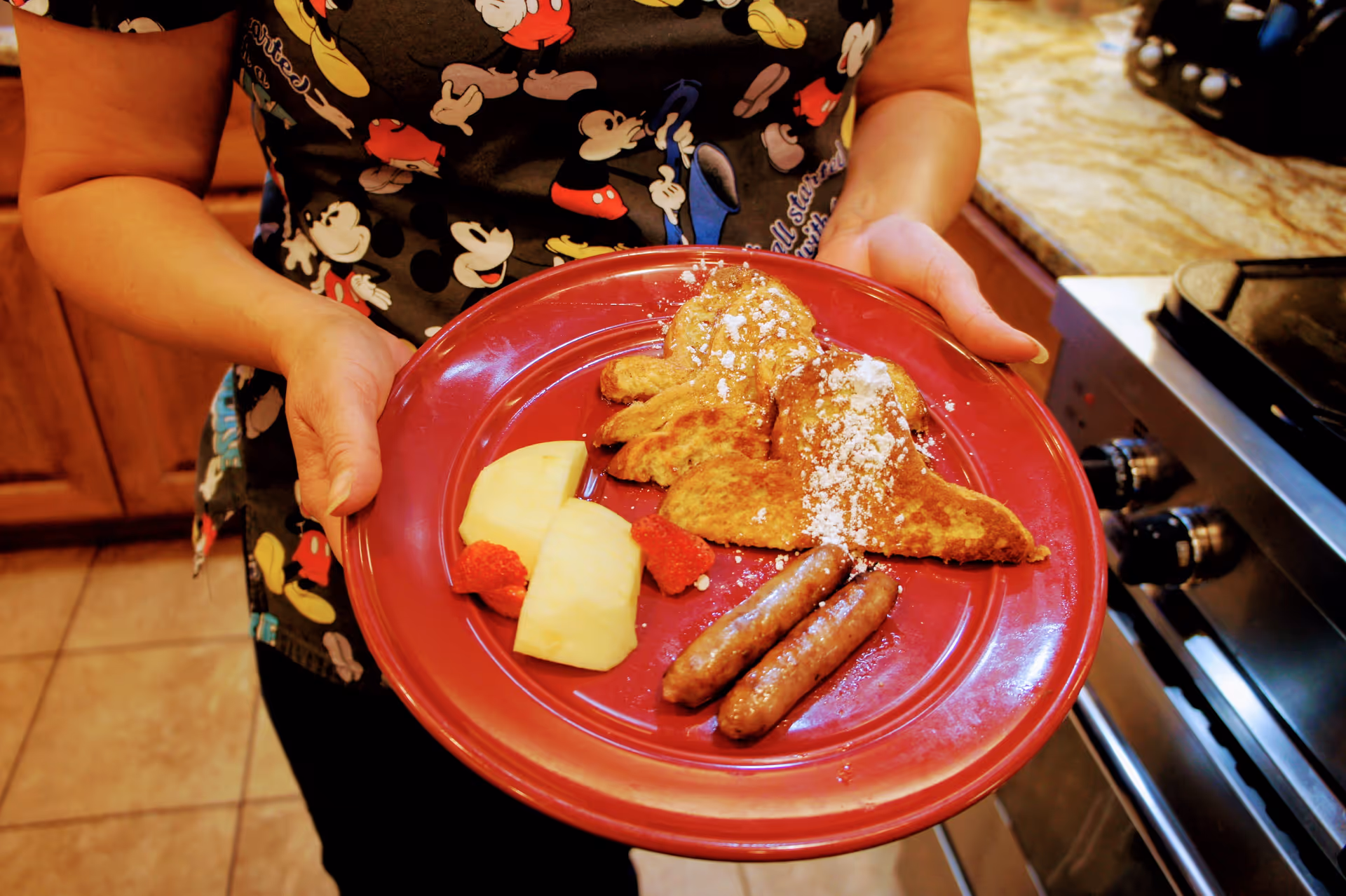 A person holds a red plate with French toast, sausages and fruit in a kitchen.