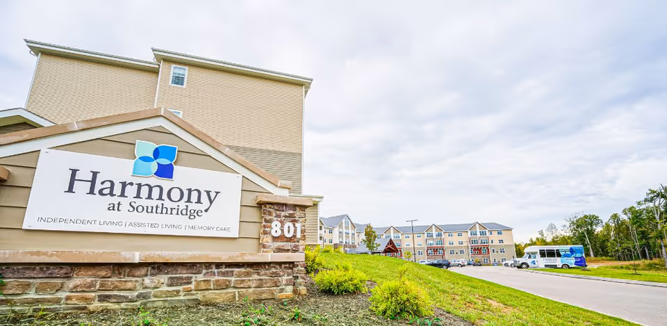 Exterior view of Harmony at Southridge senior living facility showing the main building, a large sign with the facility name and services offered, a driveway, parked cars, and a shuttle bus under a cloudy sky.