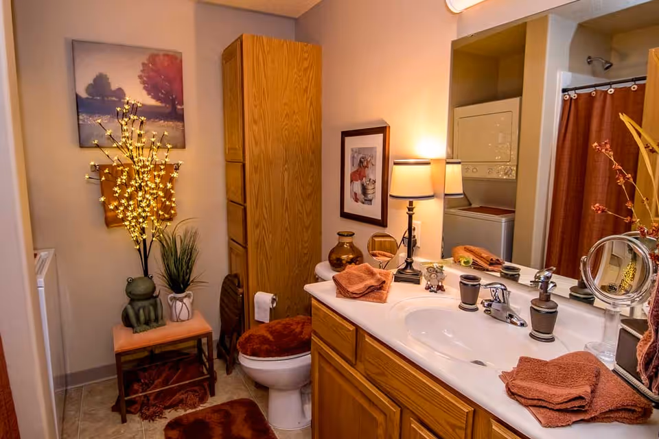A cozy bathroom with wooden cabinets and a white countertop sink. The countertop has brown towels, a small lamp, decorative items, and a mirror reflecting a stacked washer and dryer. There is a toilet with a brown seat cover, a small bench with a frog decoration, and a lit decorative branch. A shower with a brown curtain is visible in the background.