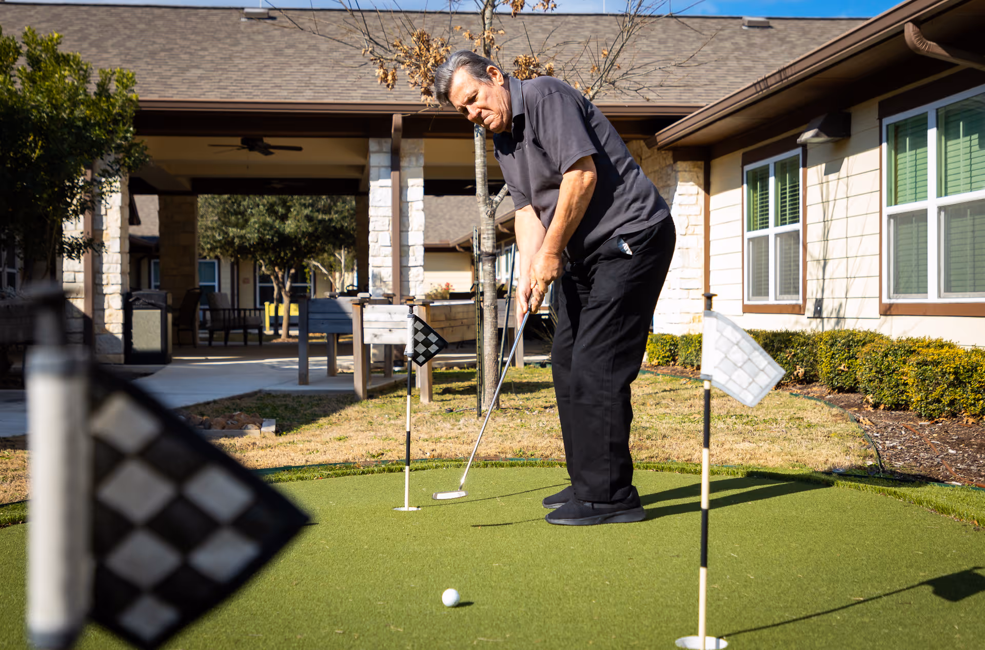 An elderly man playing putting golf on a small outdoor green at a senior living facility. The area is surrounded by buildings with windows and bushes, and there are checkered flags marking the holes on the putting green.