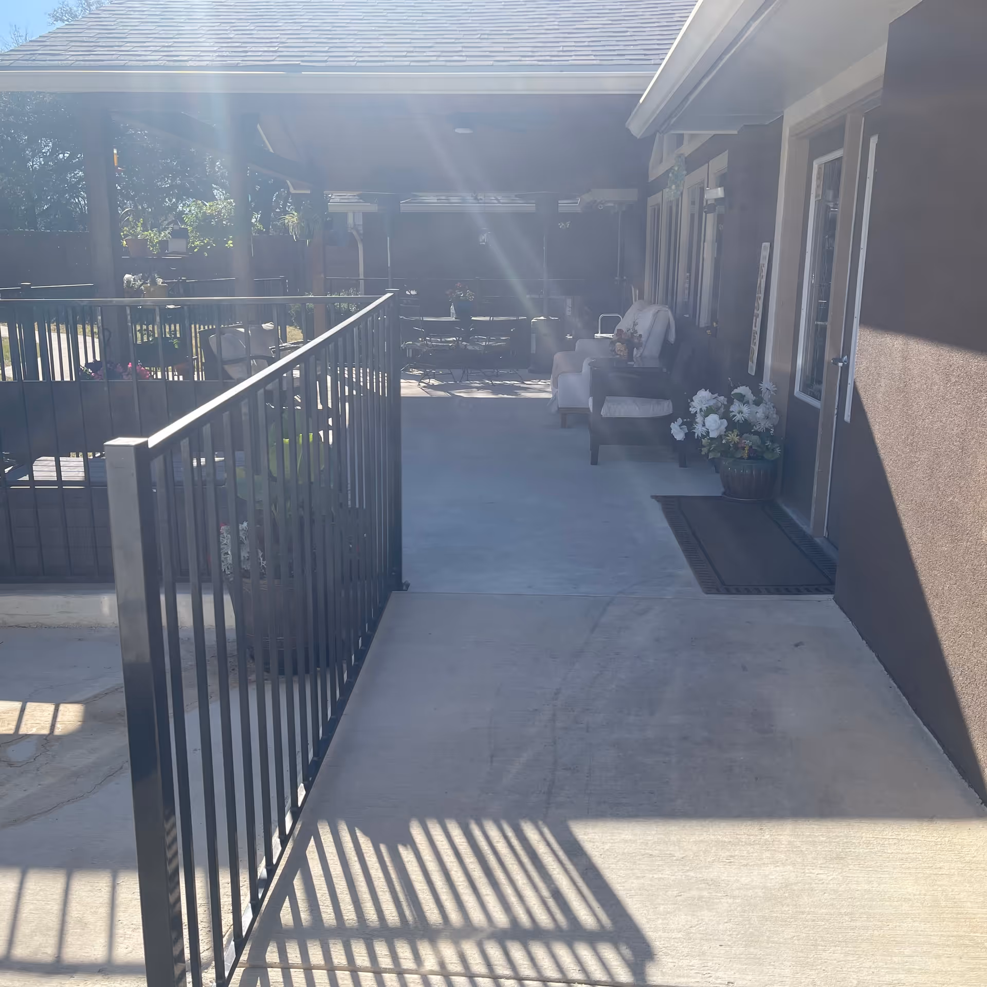 Outdoor covered patio area with seating including chairs and a small table, potted plants, and a railing along the walkway. Sunlight is shining through, casting shadows on the concrete floor.