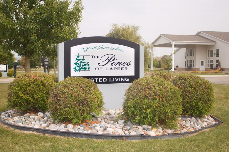 Entrance sign for The Pines of Lapeer in a rock-bordered bed with trimmed shrubs and the facility building visible behind it.