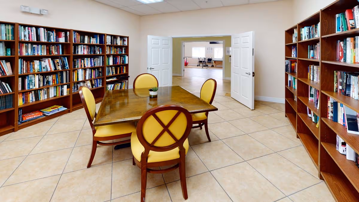 A bright interior reading room with bookshelves along the walls and a wooden table surrounded by yellow-upholstered chairs.