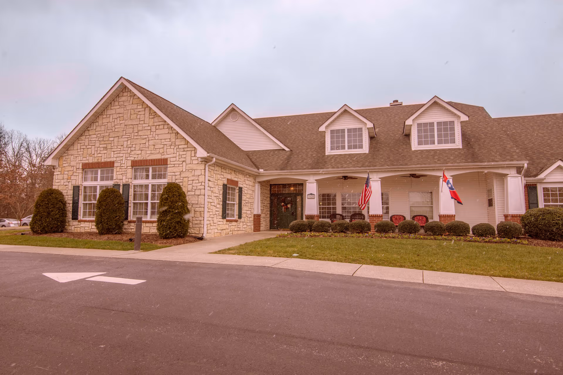 Front exterior of a one-story stone-and-siding senior living building with a covered porch, shrubs, and two flags.