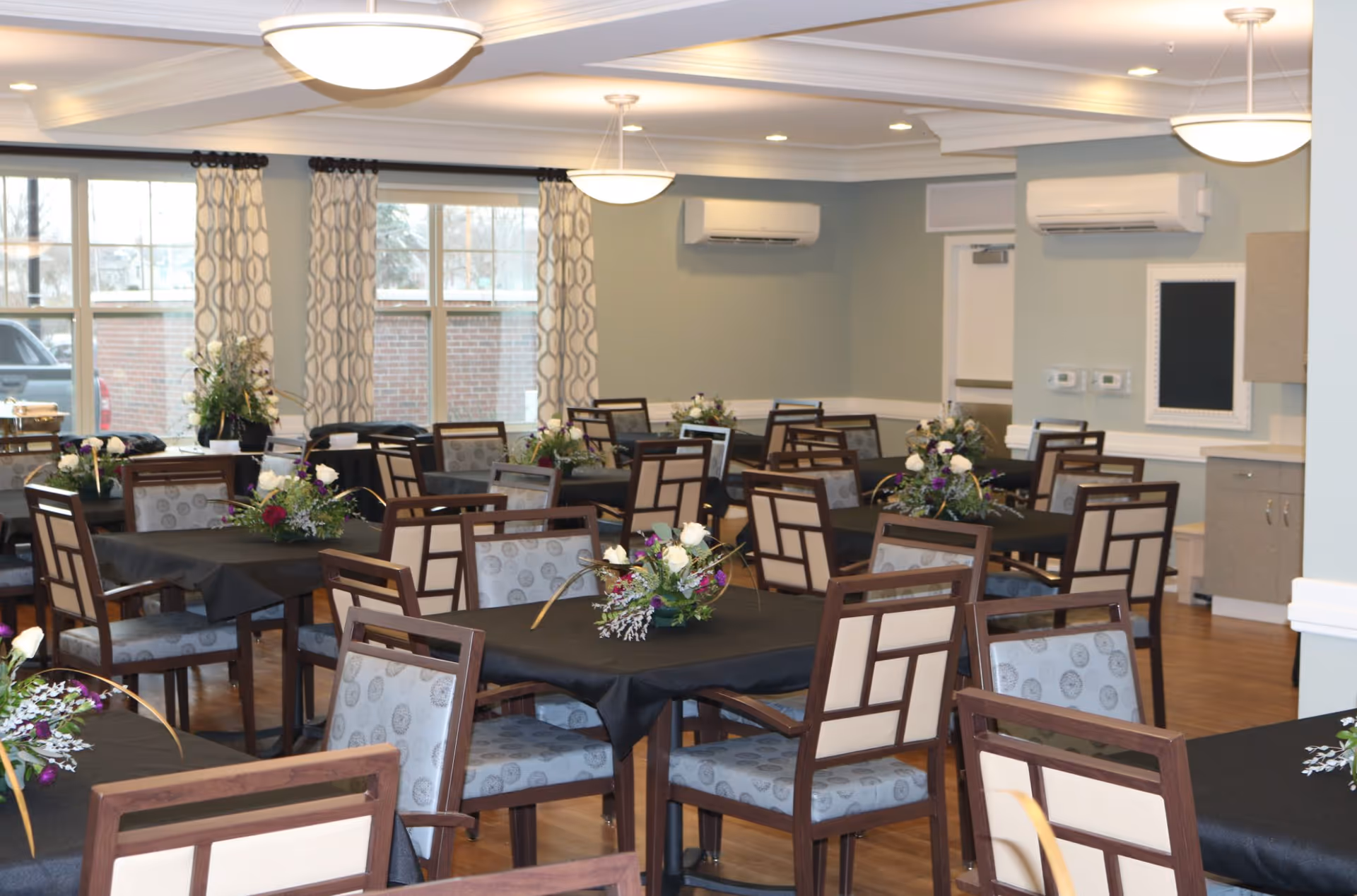 A dining room in a senior living facility with multiple tables covered in black tablecloths, each adorned with floral centerpieces. The room has wooden chairs with patterned cushions, large windows with patterned curtains, and ceiling lights providing warm illumination. The walls are painted light green with white trim, and there are air conditioning units mounted on the walls.