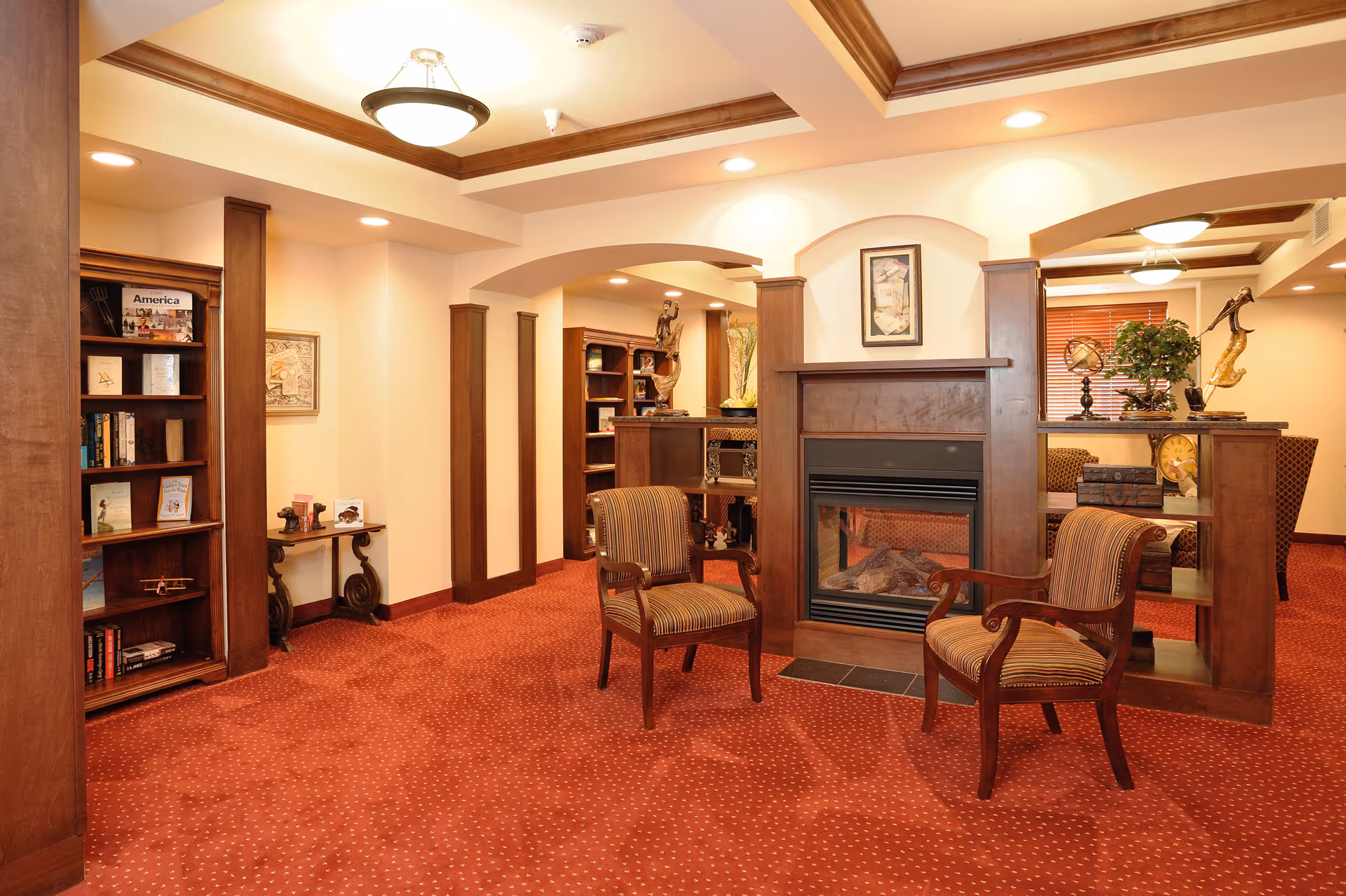 A cozy interior common area with warm lighting, featuring a fireplace surrounded by wooden paneling, two upholstered armchairs with wooden frames, bookshelves filled with books and decorative items, and a red carpet with a subtle pattern. The ceiling has recessed lighting and wooden crown molding, and there are decorative plants and artwork on the walls.