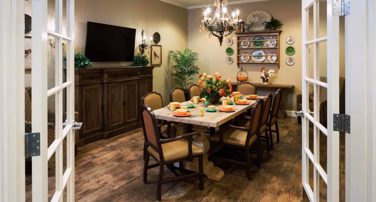 A dining room with a rectangular wooden table set for eight people with colorful plates and cups. The room features wooden flooring, a chandelier hanging above the table, a large wooden cabinet with a flat-screen TV on top, decorative plates on the wall, and a potted plant in the corner. The room is viewed through open French doors.