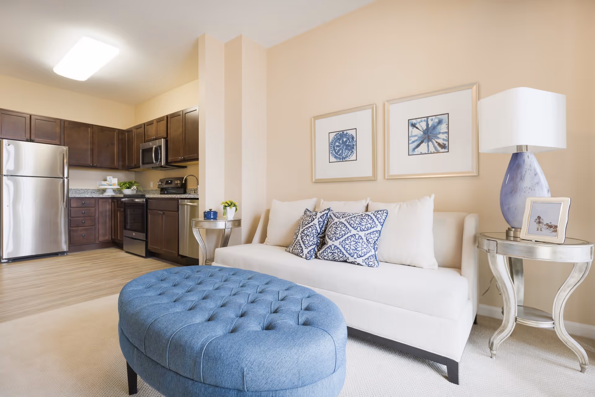 A bright and modern living area featuring a white sofa with blue and white patterned pillows, a blue tufted ottoman, and two silver side tables. One side table holds a blue lamp with a white shade and a framed photo, while the other has a small plant and a blue cup. The background shows a kitchen with dark wood cabinets, stainless steel appliances including a refrigerator, stove, microwave, and dishwasher, and granite countertops. The walls are painted a soft beige, and two framed blue and white artworks hang above the sofa.