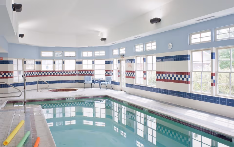 Indoor swimming pool with an adjacent hot tub in a bright tiled room lined with windows and a few chairs.