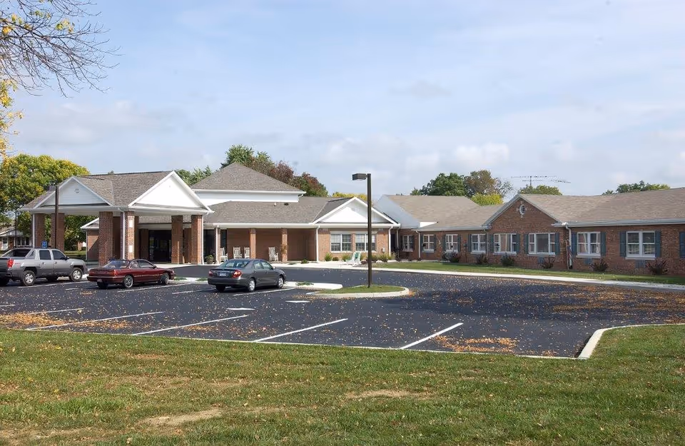 Exterior view of Grace Brethren Village showing a single-story brick building with a covered entrance and several parked cars in the parking lot. The sky is partly cloudy and there are trees in the background.