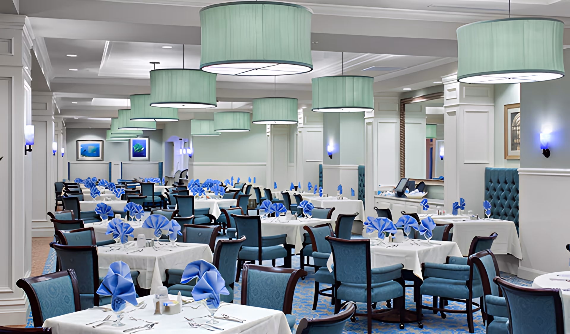 A spacious dining room with multiple tables covered in white tablecloths and neatly folded blue napkins. The room features blue upholstered chairs, large green pendant lights hanging from the ceiling, wall sconces with blue lighting, and framed artwork on the walls.