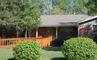 Front exterior view of a single-story brick house with a gray shingled roof, a wooden ramp leading to the front door, surrounded by green bushes and trees under a clear sky.