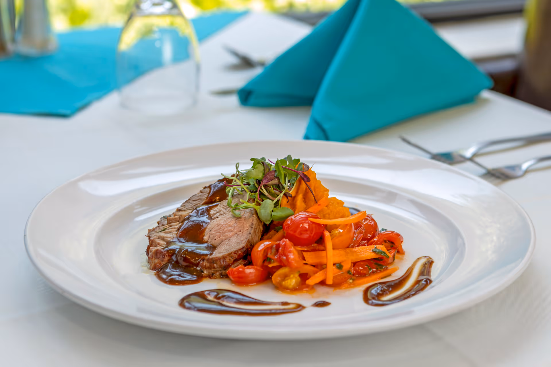 Plated gourmet roast beef with sauce, cherry tomatoes and vegetables on a white plate at a table set with teal napkins.