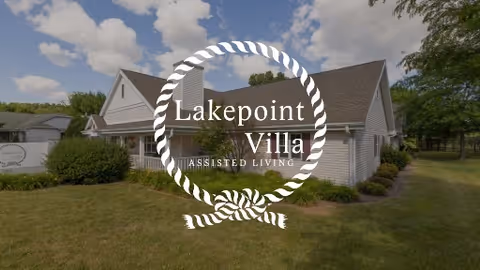Exterior view of a single-story building with white siding and a gray roof, surrounded by green grass and bushes under a partly cloudy sky. A sign for Lakepoint Villa Assisted Living is visible on the lawn.