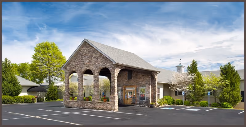 Exterior view of River Oaks Place - Lenoir City facility showing a stone entrance structure with arches, a parking lot with marked spaces including handicapped spots, and surrounding greenery under a partly cloudy sky.