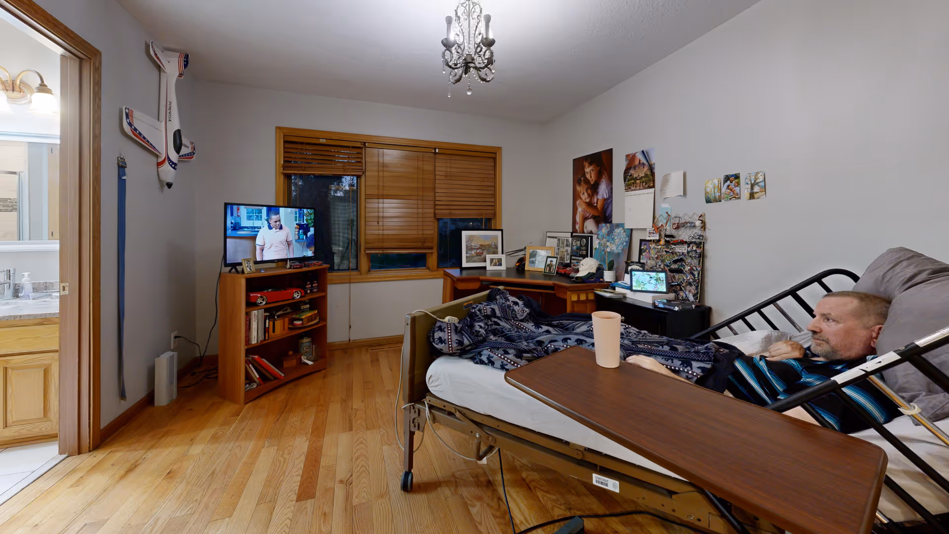A man lying in a hospital-style bed in a room with wooden floors and light gray walls. The bed has a dark brown overbed table with a cup on it. There is a wooden corner shelf with a TV showing a man in a white shirt, books, and decorative items. A window with wooden blinds is behind the shelf. The room also has a desk with framed photos, a calendar, and other personal items. A bathroom with a sink and mirror is visible through an open door on the left.