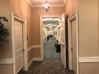 A long indoor hallway in a senior living facility with beige walls, white trim, and patterned carpet. Several closed white doors line both sides of the hallway, and a chandelier-style light fixture hangs from the ceiling. The hallway extends into the distance with additional lighting and some furniture visible at the far end.