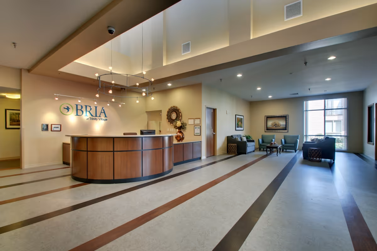 Reception area of BRIA of Trinity Village featuring a curved wooden front desk with a computer, a decorative vase with flowers, a circular chandelier above, and a seating area with sofas and armchairs near large windows letting in natural light.