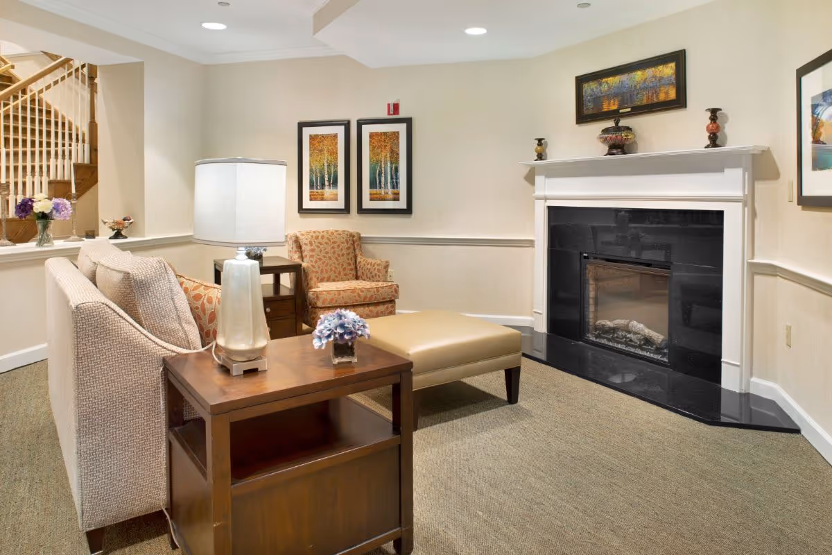A cozy living room area featuring a beige sofa with patterned cushions, a wooden side table with a lamp and a small flower arrangement, a patterned armchair, a beige ottoman, and a black fireplace with a white mantel. The walls are decorated with framed artwork, and a wooden staircase is visible in the background.