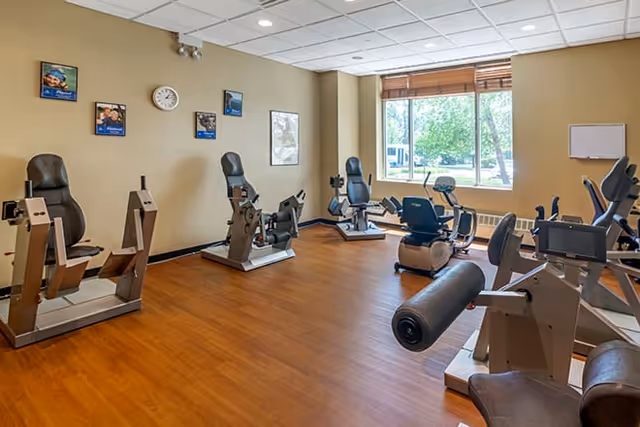 A fitness room with several exercise machines including recumbent bikes and other cardio equipment, wood-style flooring, beige walls with framed pictures, a clock, and a large window letting in natural light.