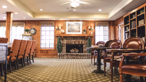 A cozy common area with a brick fireplace centered on a wood-paneled wall, flanked by two windows with blinds. The room features a patterned carpet, a row of cushioned chairs along a counter on the left, and several leather chairs around tables on the right. Decorative items include a large clock, framed artwork above the fireplace, and two small topiary trees.