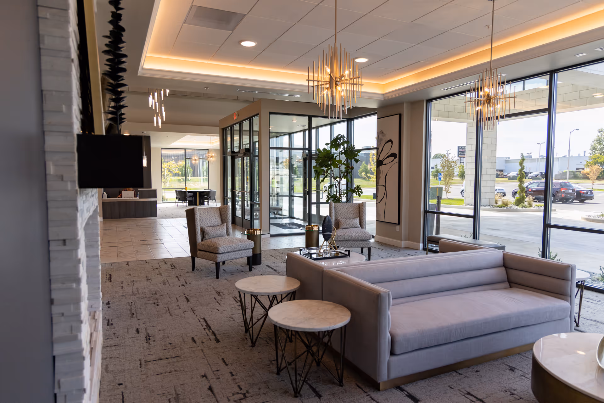 A modern and spacious lobby area with large windows allowing natural light to fill the space. The room features a light gray sofa, two armchairs, round side tables, and contemporary pendant lighting. Outside, cars and greenery are visible through the glass walls.