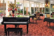 A spacious room with a black grand piano and matching bench on a red patterned carpet. The room has several tables and chairs arranged around, with large windows and glass doors letting in natural light. There is a floral arrangement on the piano and wall sconces providing additional lighting.