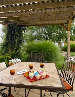 Outdoor patio area with a wooden pergola overhead, a table with a tiled surface holding a red tray with six colorful cupcakes and three glasses of iced tea with straws. Surrounding the patio are green bushes and trees.