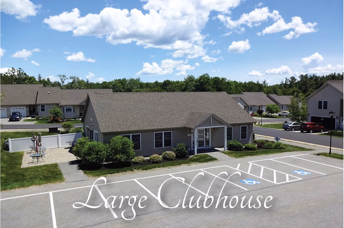 Exterior view of a single-story clubhouse building with a gray roof and beige siding, surrounded by greenery and parking spaces including handicapped spots, under a partly cloudy blue sky.