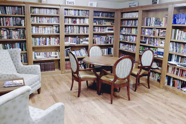 A cozy library room with wooden bookshelves filled with books lining the walls. In the center, there is a round wooden table surrounded by four upholstered chairs. Two patterned armchairs are positioned near the table, and the floor is made of light-colored wood.