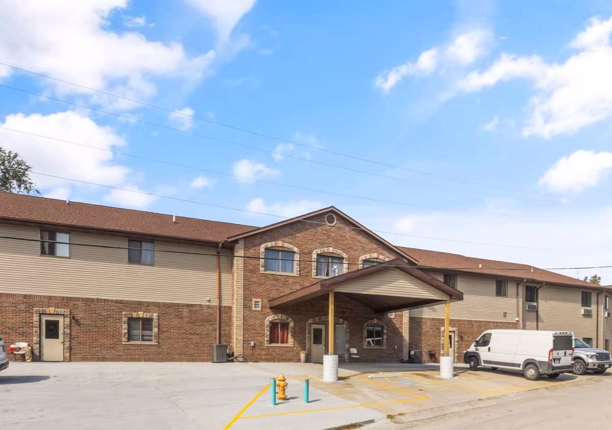 Two-story brick and siding senior living building with a covered entrance, parking area, and a white van in front.
