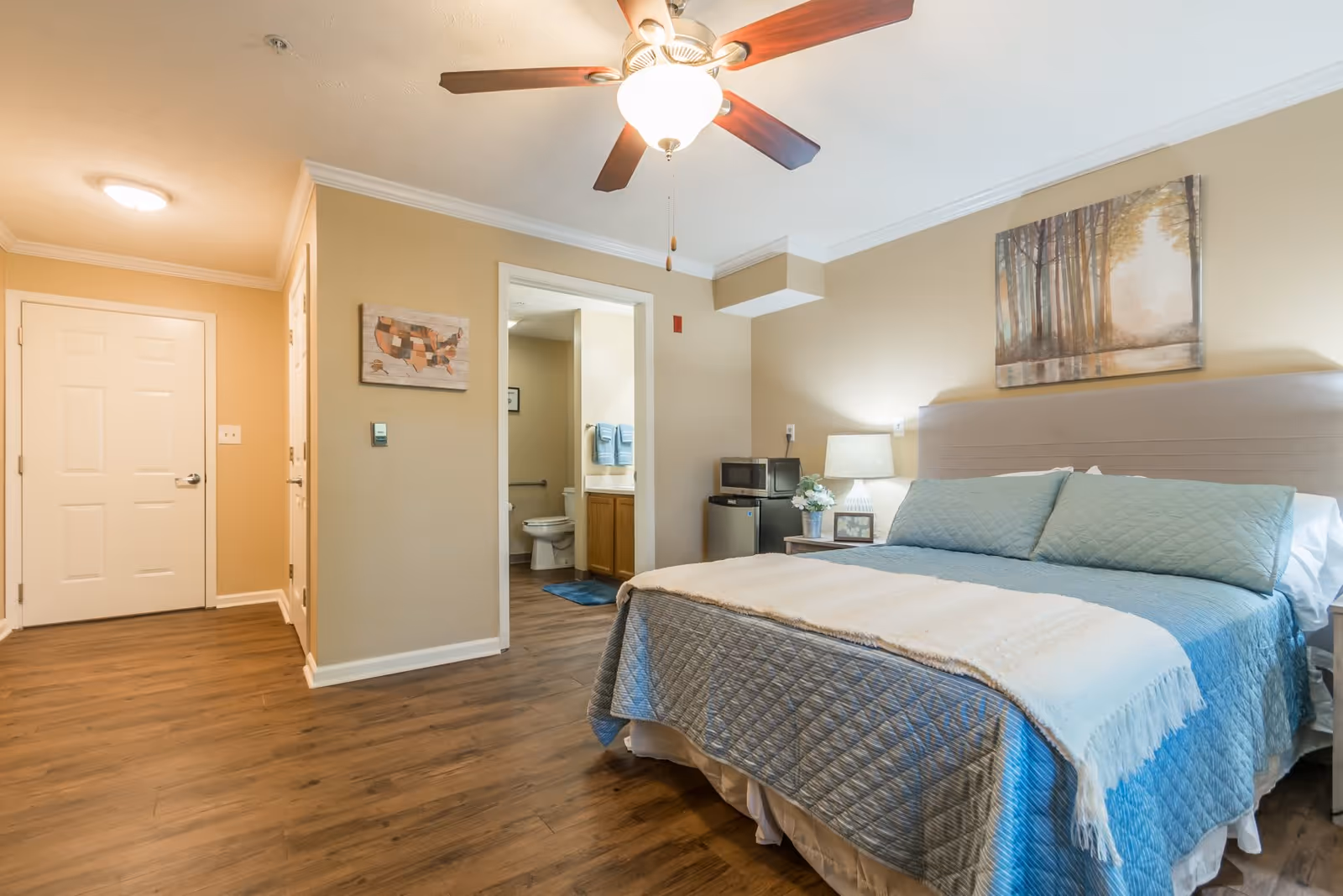 A cozy bedroom with a large bed covered in a blue quilt and a cream-colored throw blanket. Above the bed hangs a painting of a forest scene. To the right of the bed is a nightstand with a lamp, a small framed picture, and a flower arrangement. A ceiling fan with a light is mounted on the ceiling. The room has wooden flooring and beige walls. An open doorway leads to a bathroom with a visible toilet, sink, and blue towels. Near the bathroom entrance, there is a small microwave and mini-fridge.